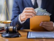 Jury Awards $15 Million in Title IX Violation Case A lawyer in a suit handles an envelope filled with cash beside a gavel and scales of justice.