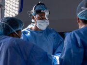 Addressing Racial Disparities in Orthopaedic Surgery Outcomes Three surgeons, dressed in blue surgical attire and face masks, are performing a medical procedure under bright surgical lights.