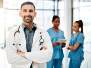 Enhance Your Orthopaedic Leadership Skills at the 2024 AOA|USC APEX A confident male doctor in a white coat stands with his arms crossed and smiles at the camera, while two nurses discuss patient care in the background.