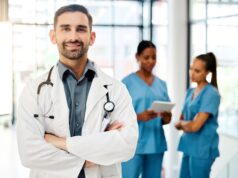 Enhance Your Orthopaedic Leadership Skills at the 2024 AOA|USC APEX A confident male doctor in a white coat stands with his arms crossed and smiles at the camera, while two nurses discuss patient care in the background.